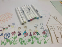 Multi-colored markers lying in a neat row on top of a child's drawing of a home and family.